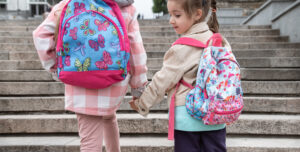 Two little girls with backpacks hold hands and go to school, view from the back.
